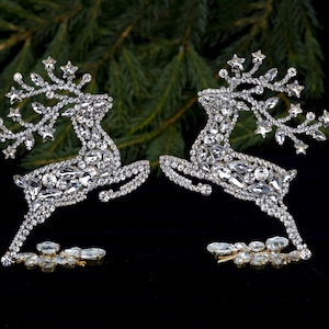 Puede incluir: Dos adornos de reno plateados cubiertos de brillantes de imitación. Los renos se miran y tienen las patas levantadas como si estuvieran saltando.