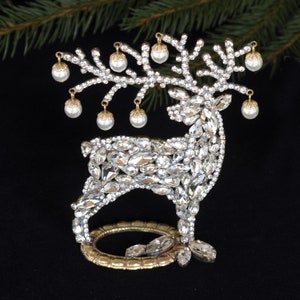 May include: A gold-tone reindeer figurine covered in clear rhinestones with a gold base. The reindeer has a branch of gold-tone leaves with white pearl-like ornaments hanging from it.