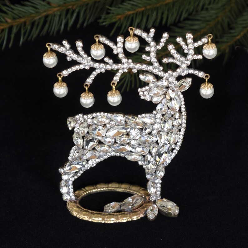 May include: A gold-tone reindeer figurine covered in clear rhinestones with a gold base. The reindeer has a branch of gold-tone leaves with white pearl-like ornaments hanging from it.