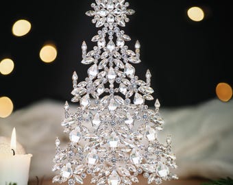 May include: A decorative crystal Christmas tree, featuring a star at the top and numerous clear, faceted crystals. It stands on a gold filigree base. The background is dark with blurred lights, and a lit candle is visible.