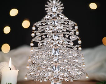 May include: A decorative Christmas tree made of clear crystals and faux pearls. The tree has a gold-colored base and is adorned with various sizes of sparkling crystals. Small faux pearls hang from the branches. A lit candle is in the foreground.