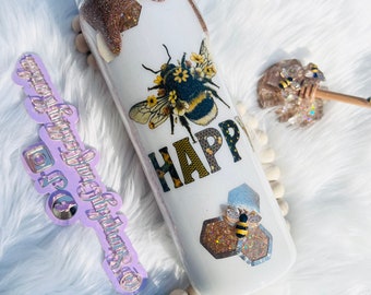May include: White tumbler with a gold glitter rim and a bee with flowers design. The tumbler has the word "HAPPY" written on it. There is a smaller bee on a honeycomb design on the bottom of the tumbler.