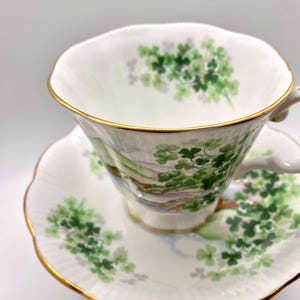 Puede incluir: Un juego de taza y platillo blancos con borde dorado. La taza y el platillo están decorados con tréboles verdes y un paisaje. La taza tiene un asa curvada.