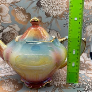 Puede incluir: Una tetera de cerámica colorida con un esmalte de arcoíris, ribetes dorados y un adorno dorado en la tapa. La tetera mide aproximadamente 15 cm de altura. El fondo presenta un estampado floral.