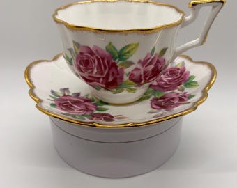 Puede incluir: Juego de taza y platillo de té blanco con un diseño floral de rosas rosas y verdes. Los bordes festoneados y dorados añaden elegancia. La taza descansa sobre el platillo.