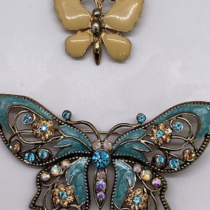 Puede incluir: Dos broches de mariposa, uno es una mariposa simple de oro y crema, el otro es una mariposa más adornada de azul y oro con pedrería.