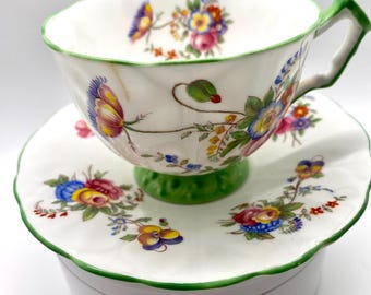 Könnte beinhalten: Ein weißes Tee-Service mit einer Tasse und Untertasse mit floralem Muster. Die Tasse hat einen grünen Henkel und Fuß, sowie einen grünen Rand. Die Untertasse hat ein passendes Blumenmuster und einen grünen Rand. Das Set steht auf einer weißen Oberfläche.