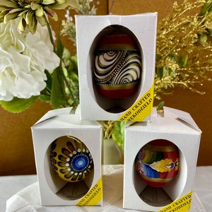May include: Three hand-painted Easter eggs in individual boxes. The eggs are decorated with intricate patterns in gold, red, black, and white. The boxes are white with yellow labels that read "HAND CRAFTED RĘKODZIEŁO".
