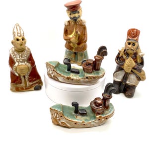 May include: Set of five ceramic figurines, including three figures of men in military uniforms and two small boats. The figures are painted in shades of brown, red, and green. The boats are painted in shades of green and brown.
