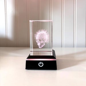 May include: A clear, rectangular glass block with a 3D image of a skull with a spiky crown. The block sits on a black base with a pink light and a power button symbol.