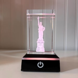 May include: A clear, rectangular glass block featuring a 3D rendering of the Statue of Liberty, illuminated in pink. The statue sits on a black base with a power button and a pink light strip.