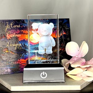 May include: A lighted glass block featuring a 3D teddy bear design with the words "I Love You". The block sits on a gray base with an on/off power button. A canvas with the text "Ja Cara Custom Designs" is in the background.
