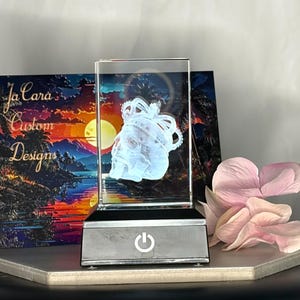 May include: A rectangular glass block with a skull design and crown, illuminated by a light. The base is silver with a power button. A colorful landscape painting and pink flower petals are in the background. The text "Ja Cara Custom Designs" is visible.