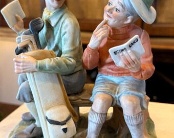 May include: A porcelain figurine of two golfers sitting on a bench. The male figure wears a light green shirt and cap, holding a golf bag. The female figure wears a coral sweater, blue shorts, and a white hat, holding a scorecard labeled SCORE CARD.