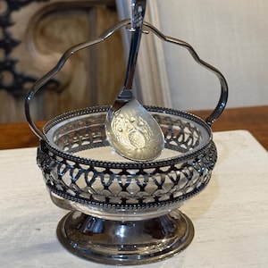 May include: A silver-plated sugar bowl with a clear glass insert and a matching spoon. The bowl has a decorative openwork design around the rim and a handle. The spoon rests inside the bowl.