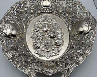 May include: Ornate silver-toned serving tray with a floral centerpiece. The tray features intricate openwork detailing around the edges and handles. The center of the tray has a raised floral design. The tray is likely made of metal.
