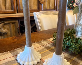May include: Two white glass candle holders with a scalloped design and brown candles. The candle holders are on a plaid tablecloth. The candles are a light brown color.