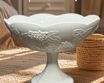 May include: A white, pedestal fruit bowl with a scalloped rim and grape vine detailing. The words "JUST IN!" are displayed above the bowl. The bowl is on a woven mat, with a woven basket in the background.