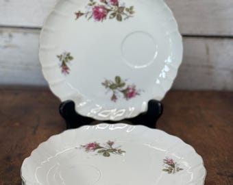 May include: A stack of white porcelain plates with a scalloped edge and a floral rose design in pink and green. The plates have a small circular indentation in the center. One plate is displayed on a black stand.