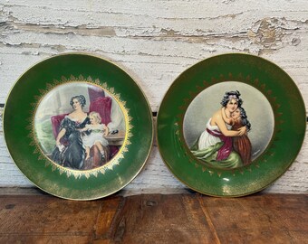 May include: Two decorative plates with green rims and gold accents. Each plate features a central portrait of women and children in a classical style. The plates are displayed on a wooden surface.