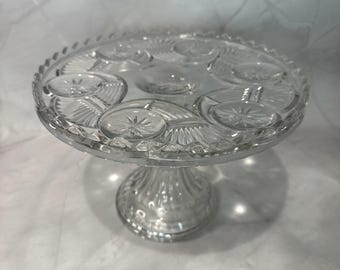 May include: A clear glass cake stand with a round, decorative top and a pedestal base. The top features a starburst and oval design. The stand is made of clear glass and is suitable for displaying cakes or other desserts.