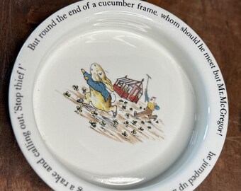May include: White ceramic plate featuring a colorful illustration of Peter Rabbit running from Mr. McGregor. The plate has text around the rim that reads, "But round the end of a cucumber frame, whom should he meet but Mr. McGregor!"