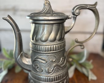 May include: Antique silver-toned metal coffee pot with a lid and curved handle. The pot features ornate detailing, including textured patterns and a decorative handle. The spout is curved, and the lid has a small finial. The pot is likely vintage.