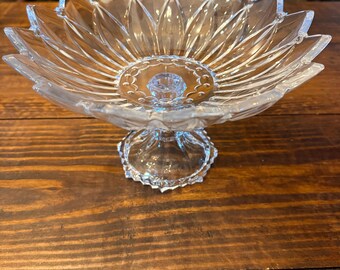 May include: A clear glass pedestal bowl featuring a scalloped, petal-like rim and a circular base. The interior of the bowl displays a textured pattern of radiating lines and a central medallion design with small circular indentations.