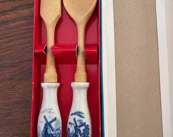 May include: A wooden fork and spoon set with white ceramic handles featuring a blue and white windmill illustration. The utensils are presented in a red cardboard box with a separate white lid, showcasing a classic design with light wood heads.