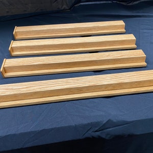 May include: Four light-colored wooden Scrabble tile holders. Each holder is rectangular with a recessed channel to hold tiles. The wood grain is visible. The holders are arranged in a staggered pattern on a dark blue surface.