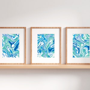 Pu&ograve; includere: Tre stampe incorniciate con disegni astratti marmorei blu e verdi. Le stampe sono esposte su uno scaffale di legno contro una parete bianca.