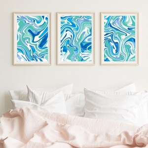 Pu&ograve; includere: Tre stampe d'arte astratta incorniciate con un designo marmoreo blu, verde e bianco. Le stampe sono appese a un muro bianco sopra un letto con cuscini bianchi e una coperta rosa.