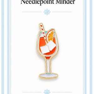 Könnte beinhalten: Ein Emaille-Nadelmagnet in Form eines goldgeränderten Cocktailglases mit orangefarbener Flüssigkeit, zwei weißen Eiswürfeln, einer Orangenscheibe und einem rot-weiß gestreiften Strohhalm. Der Text Needlepoint Minder und 2coshop ist sichtbar.