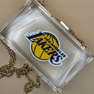 May include: A clear acrylic clutch bag with a gold chain strap. The bag features a Los Angeles Lakers logo patch with a basketball and the team name in purple and yellow.