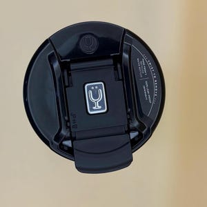 Op de afbeelding: Witte keramische mok met een zwarte handgreep en de tekst "But First, Coffee" in zwarte letters. De mok staat tegen een vage achtergrond, wat een gezellige omgeving suggereert.