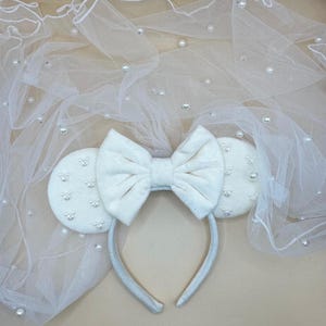 May include: A white headband with mouse ears and a large bow, decorated with small pearls. A sheer white veil with scattered pearls is draped behind the headband, creating a whimsical and elegant accessory.