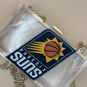 May include: A clear acrylic clutch with a gold clasp and chain strap. A navy blue patch with the words "PHOENIX SUNS" in white and a basketball design is on the clutch.