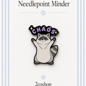 May include: A needlepoint minder enamel pin featuring a light gray and white raccoon with its arms raised. Above the raccoon, a black banner with purple text reads CHAOS, accented by two small yellow stars. The pin has a black metal outline.