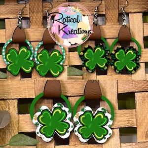 May include: Four pairs of green shamrock earrings with gold accents. Each pair has a brown leather strap and a green circle. The earrings are displayed on a woven wicker background.