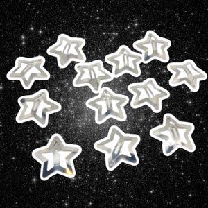 May include: A collection of silver star-shaped hair clips with white outlines, scattered against a black background with sparkling glitter. The clips are made of metal and have a simple, elegant design.