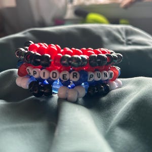May include: A stack of colorful beaded bracelets. The bracelets are red, black, white, and blue. One bracelet spells out "SPIDER PUNK" in white letter beads. The bracelets are stacked on top of each other.