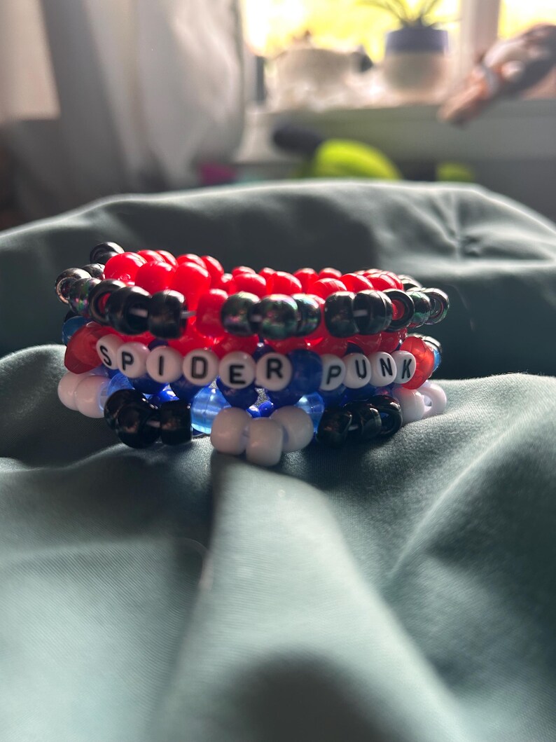 May include: A stack of colorful beaded bracelets. The bracelets are red, black, white, and blue. One bracelet spells out "SPIDER PUNK" in white letter beads. The bracelets are stacked on top of each other.