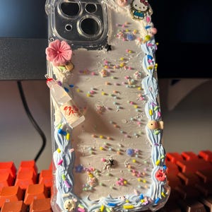 Puede incluir: Funda de tel&eacute;fono transparente decorada con una flor rosa, una botella en miniatura y un charm de Hello Kitty. La funda presenta un borde azul claro con chispas y adornos coloridos en forma de estrella.