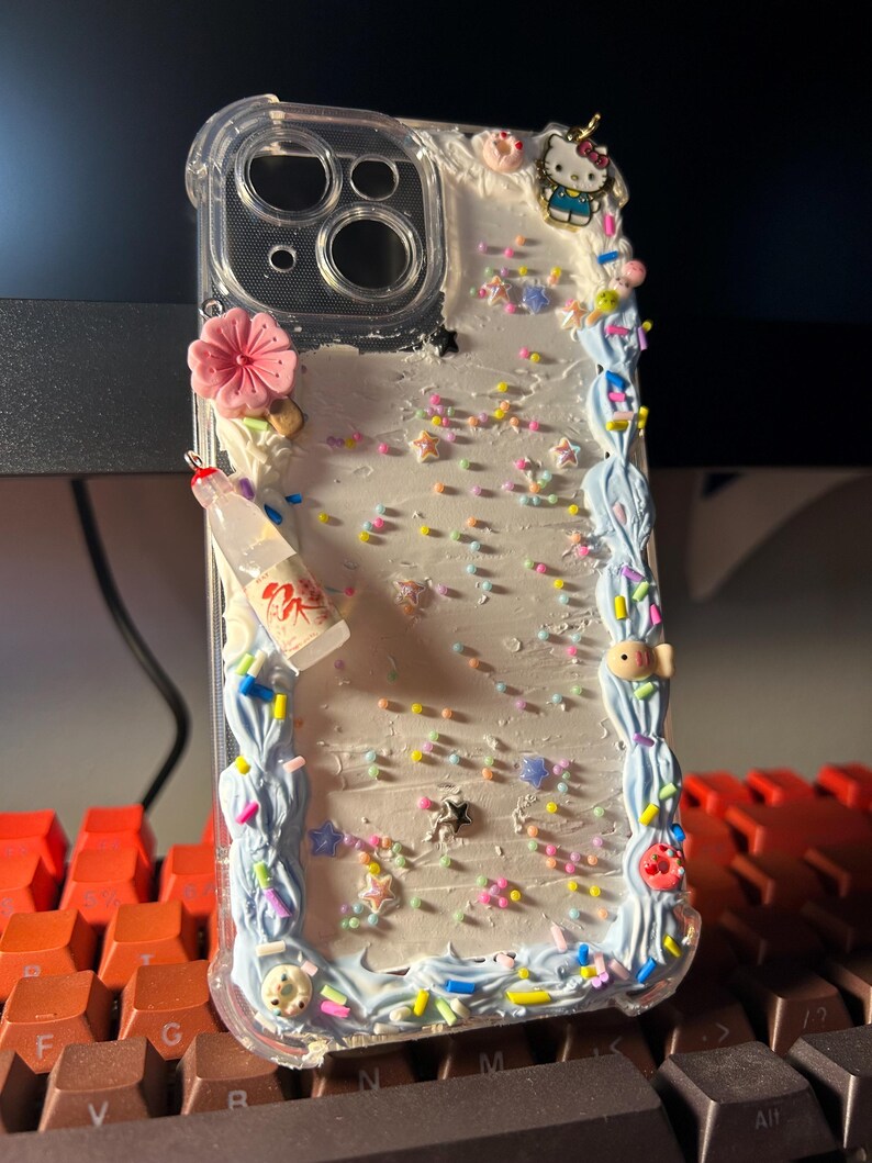 Puede incluir: Funda de tel&eacute;fono transparente decorada con una flor rosa, una botella en miniatura y un charm de Hello Kitty. La funda presenta un borde azul claro con chispas y adornos coloridos en forma de estrella.