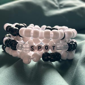 May include: A stack of three beaded bracelets. The bracelets are made of white and black beads. The center bracelet spells out the word "SPOT" with white letter beads. The bracelets are on a teal surface.