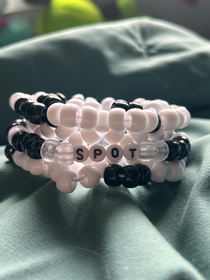 May include: A stack of three beaded bracelets. The bracelets are made of white and black beads. The center bracelet spells out the word "SPOT" with white letter beads. The bracelets are on a teal surface.