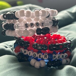 May include: Two stacked beaded bracelets. The top bracelet is white and black with the word "SPOT" spelled out. The bottom bracelet is red, black, blue, and white with the words "SPIDER PUNK".