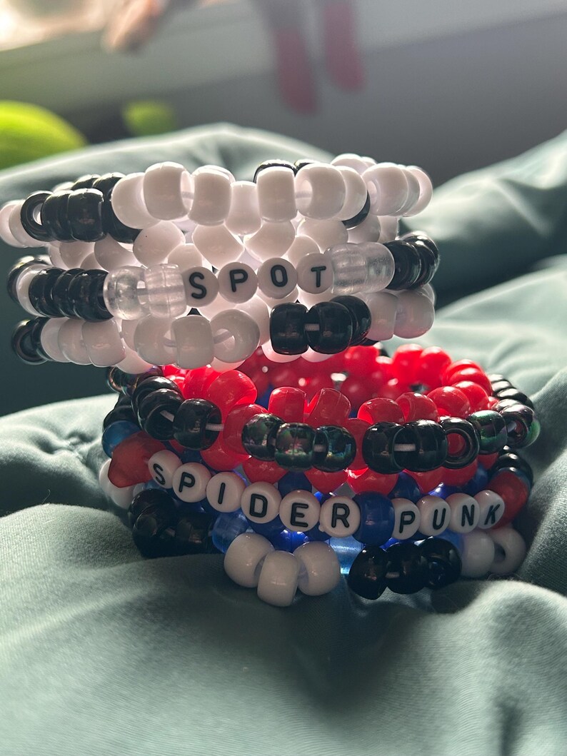 May include: Two stacked beaded bracelets. The top bracelet is white and black with the word "SPOT" spelled out. The bottom bracelet is red, black, blue, and white with the words "SPIDER PUNK".