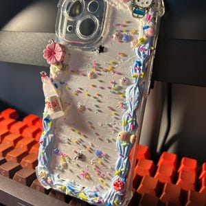 Puede incluir: Funda de tel&eacute;fono transparente con un dise&ntilde;o decorativo. La funda presenta un borde de olas azules, chispas de colores y peque&ntilde;os amuletos, incluyendo una flor rosa, un amuleto de Hello Kitty y una botella en miniatura.
