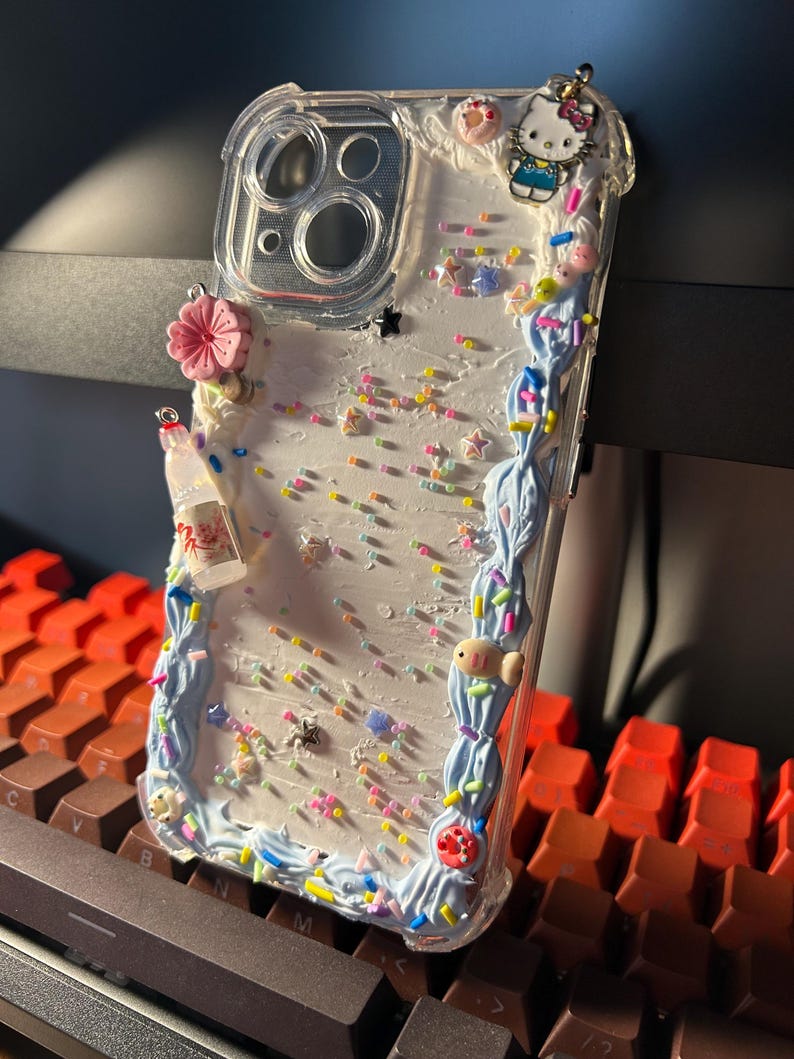 Puede incluir: Funda de tel&eacute;fono transparente con un dise&ntilde;o decorativo. La funda presenta un borde de olas azules, chispas de colores y peque&ntilde;os amuletos, incluyendo una flor rosa, un amuleto de Hello Kitty y una botella en miniatura.